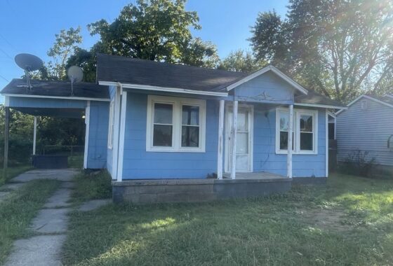 Small blue house with white trim and a carport on a sunny day.