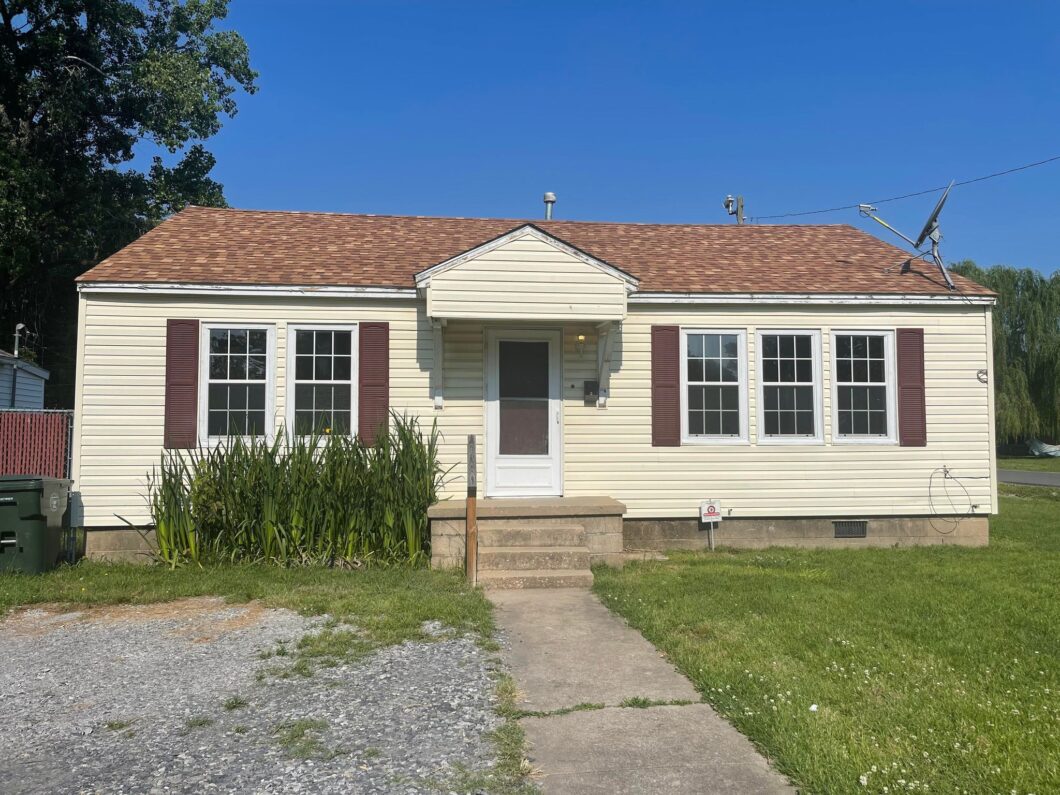 A small beige house with red shutters and a tiled roof on a sunny day.